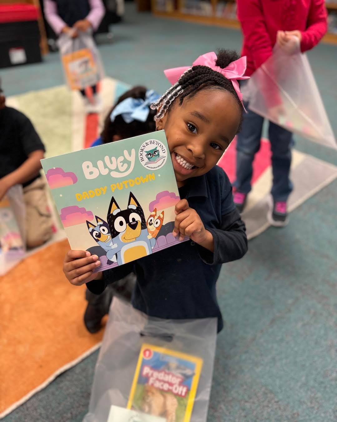 A smiling young reader with a pink bow holding a Bluey book