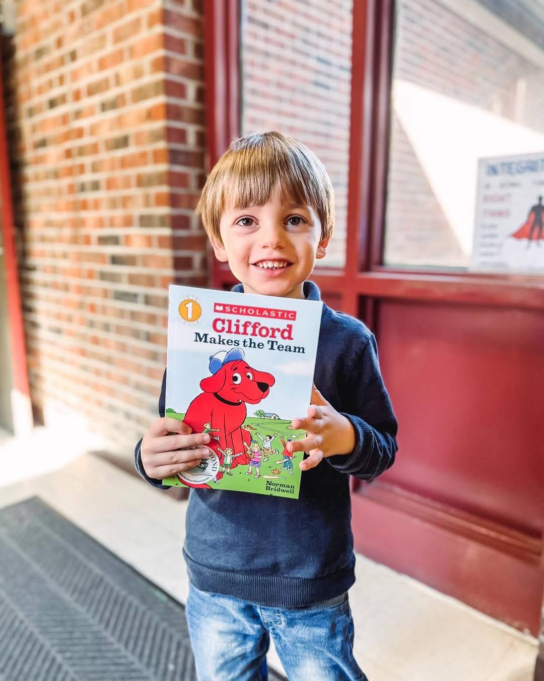 A smiling young reader holding Clifford Makes the Team at school